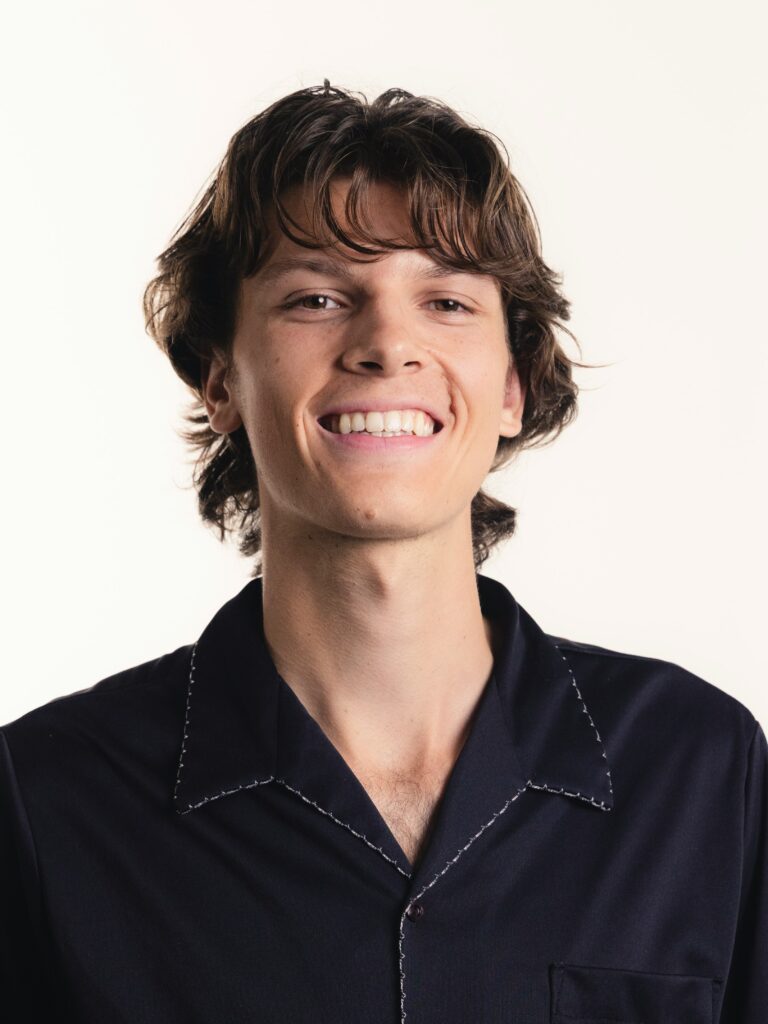 [ALT_TEXT: Un joven con un corte mullet con capas y textura, vestido con camisa azul oscuro, sonriendo.]