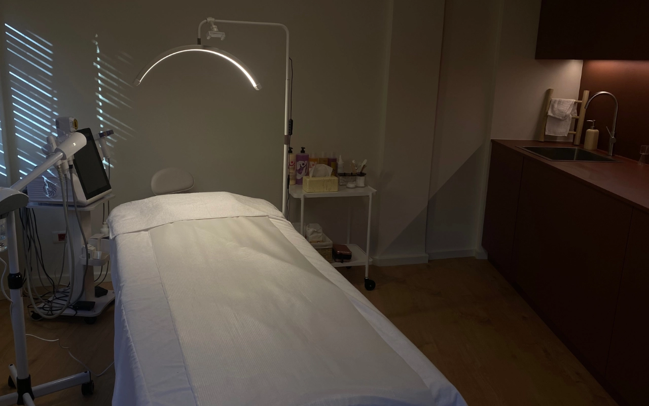 Una sala de tratamiento enfocada con una cama blanca bajo una lámpara LED de arco, equipo profesional de estética con pantalla digital, productos para el cuidado de la piel en un carrito y armarios de madera oscura con lavabo a lo largo de la pared del fondo.