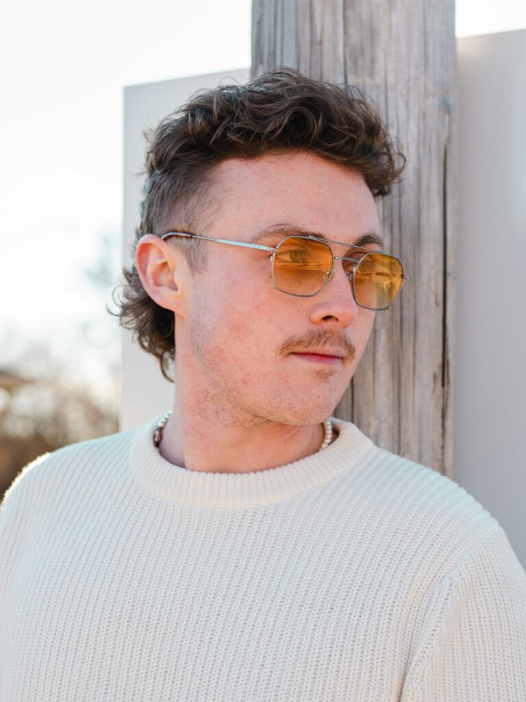 [ALT_TEXT: Un joven con un corte mullet rizado, apoyado en una pared al aire libre en un día soleado.]