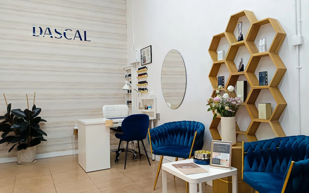 Un interior de salón de belleza elegante con sillones de terciopelo azul oscuro y una estantería de pared hexagonal de madera. Una mesa de manicura blanca se sita en la esquina contra una pared texturizada de color claro con la palabra "DASCAL" en letras azules.