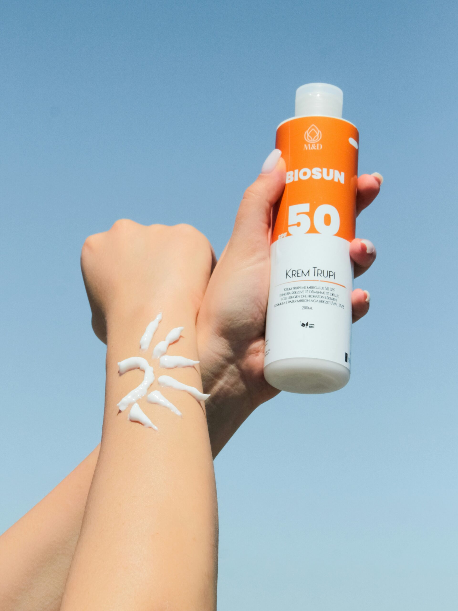 Primer plano de los brazos de una mujer contra un cielo azul, una mano sostiene un bote de protector solar y en un antebrazo tiene crema solar con forma de sol