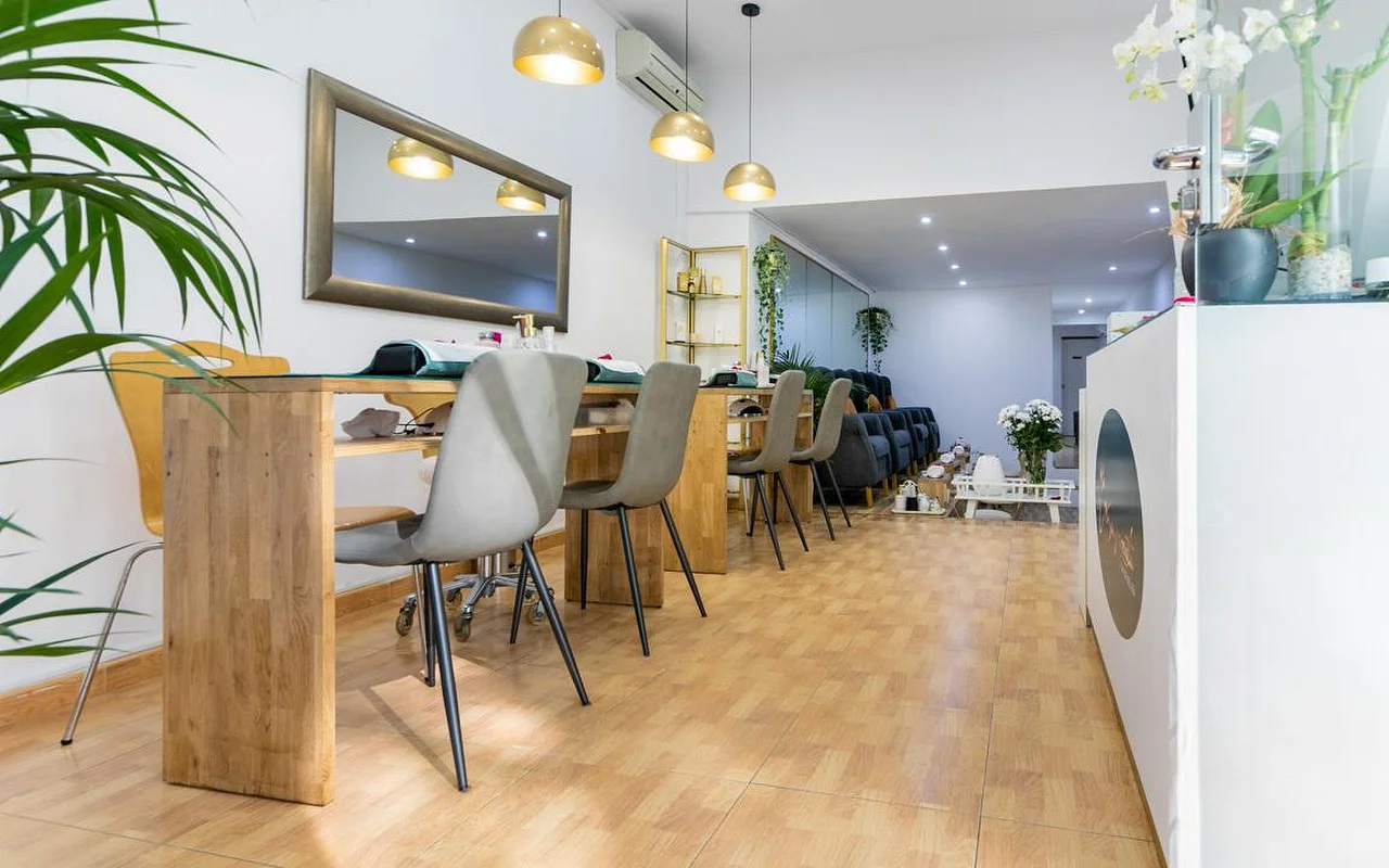 Un salón de uñas luminoso con largas mesas de manicura de madera y sillas modernas grises. Lmparas colgantes doradas de forma redondeada iluminan las estaciones, y un gran espejo con marco oscuro cuelga de la pared blanca.