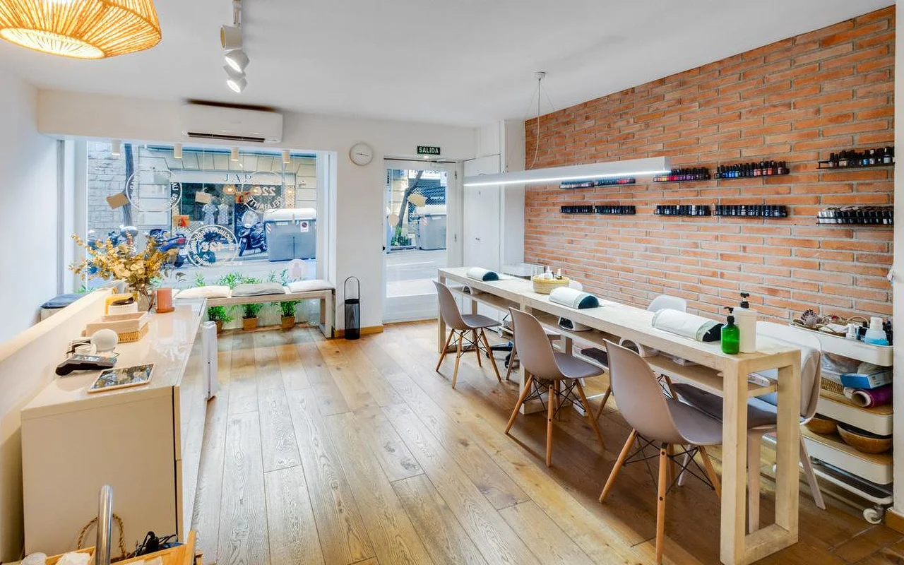 Un salón contemporneo con un toque rstico, con una larga barra de manicura blanca y una pared de ladrillo rojo visto. Grandes ventanales dan a una calle de la ciudad, llenando de luz natural el espacio de suelo de madera.