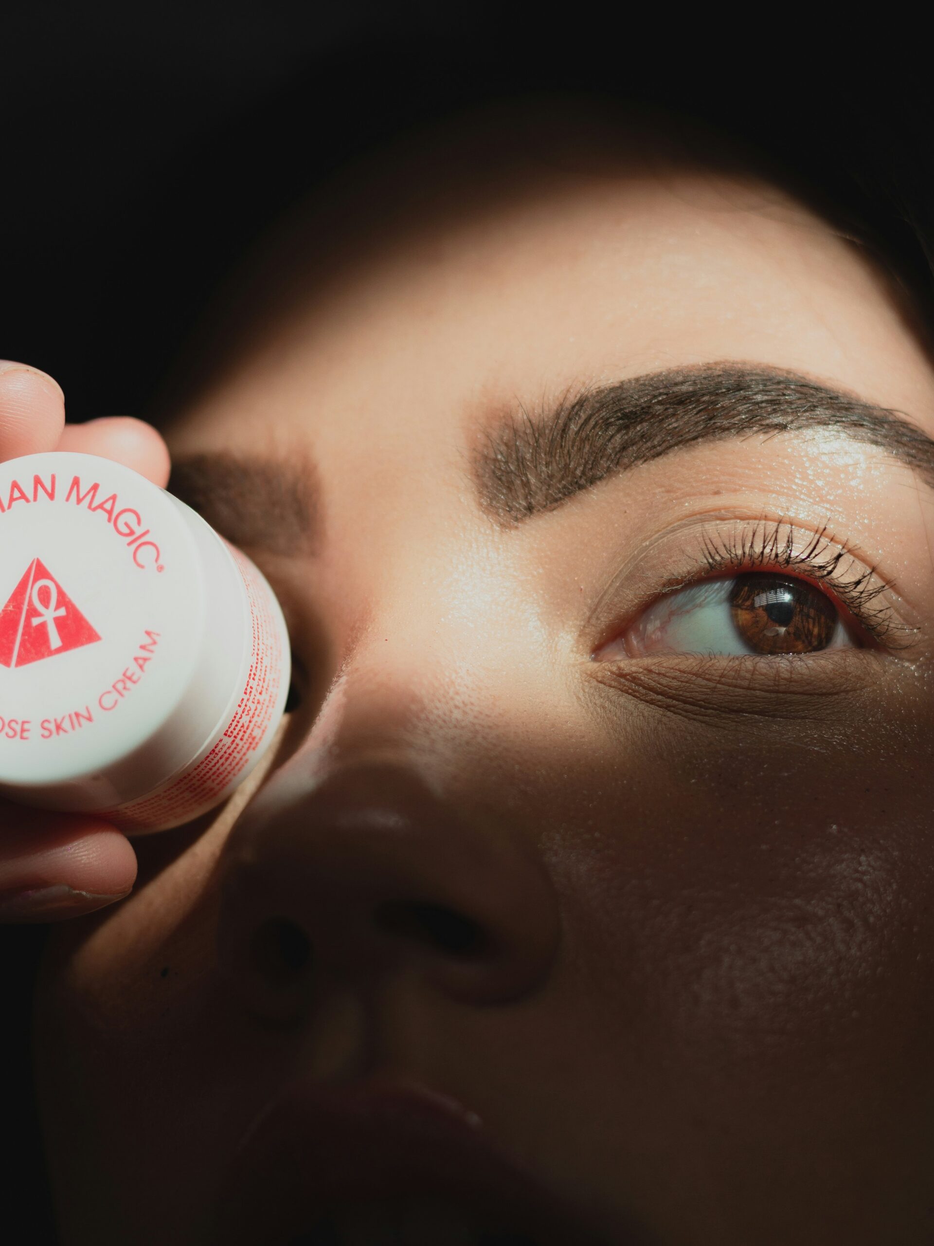 Primer plano del rostro de una mujer con microblading en las cejas sosteniendo un pequeño tarro de crema delante de un ojo