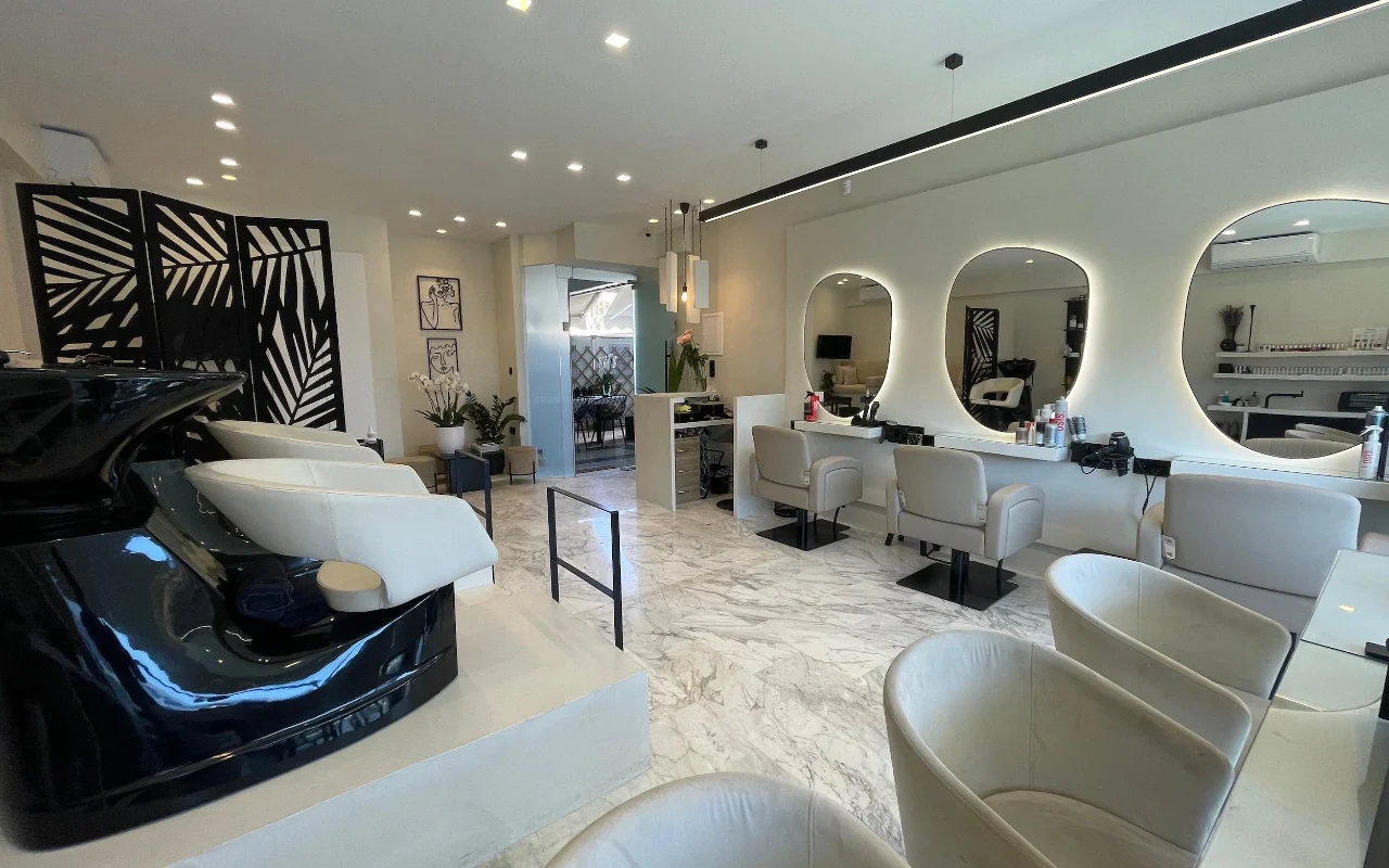 Una peluquería elegante con suelos de mármol blanco, sillones de estilismo en cuero crema bajo espejos ovalados con iluminación LED perimetral, una estación de lavado en negro brillante y un biombo geométrico con motivos de hoja de palmera.