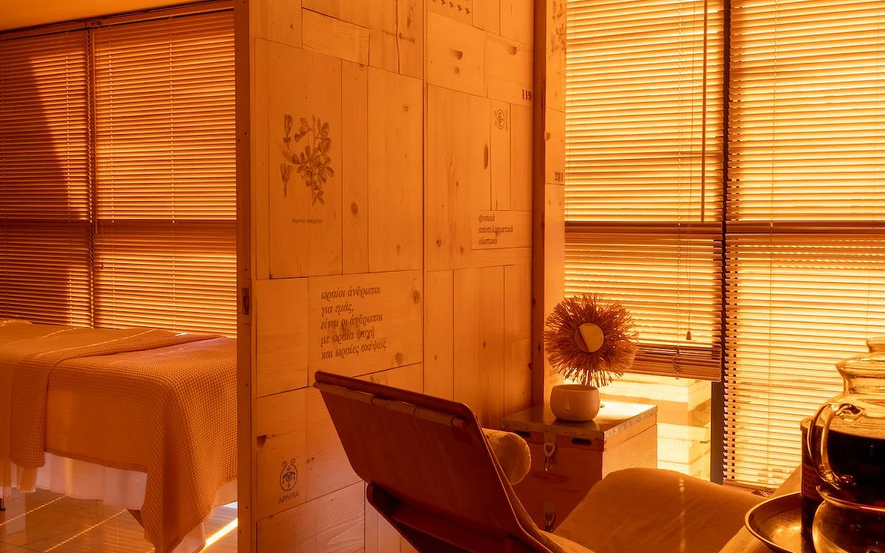 Una sala de tratamiento de spa con iluminación cálida ámbar, paneles de madera natural, ilustraciones botánicas y texto en griego grabado en la pared, una camilla de tratamiento cubierta con una manta de tejido waffle y el logotipo de APIVITA en la esquina.