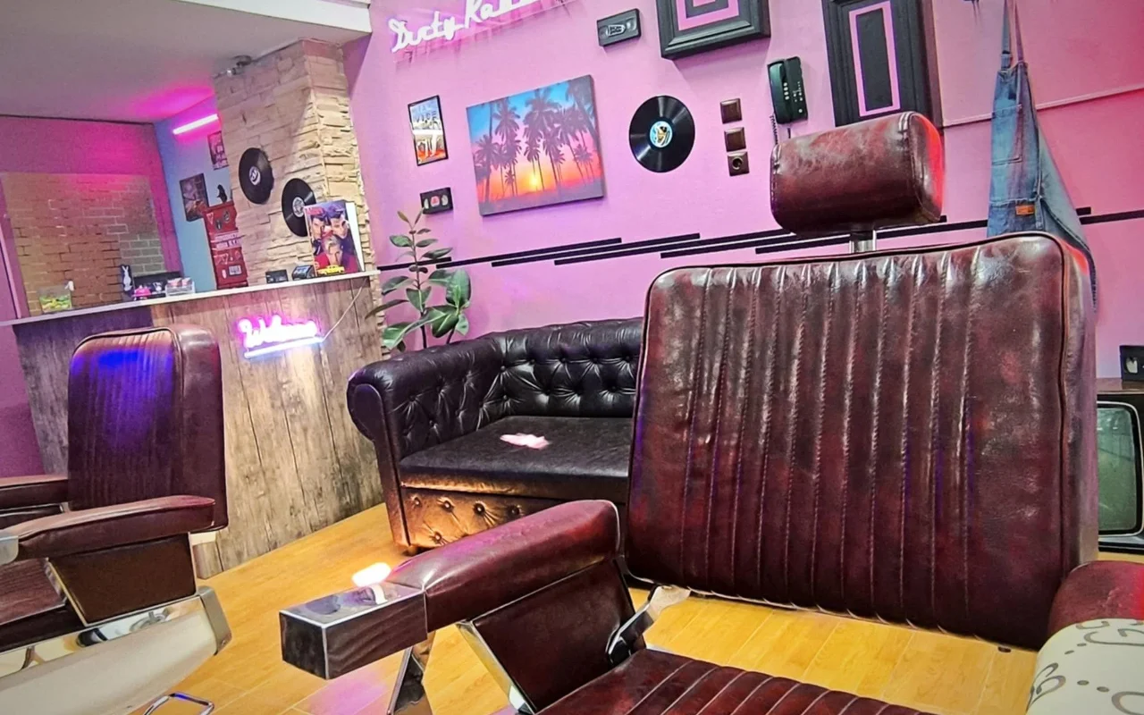 Una barbería de estilo retro con paredes rosas y moradas, letreros de neón, discos de vinilo como decoración, una pintura de una puesta de sol con palmera, sillones de barbero en cuero oscuro y un sofá Chesterfield sobre suelo de madera cálida.