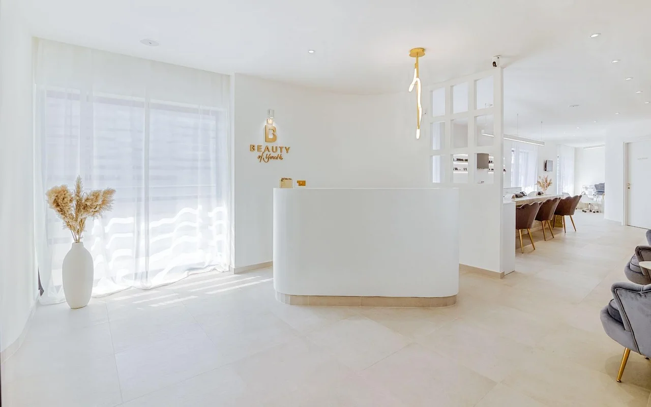 Una recepción luminosa y completamente blanca con un mostrador curvado, el logotipo dorado de Beauty of Youth en la pared, una lámpara colgante escultórica, cortinas translúcidas filtrando la luz natural y sillones de tratamiento en terciopelo marrón visibles al fondo.