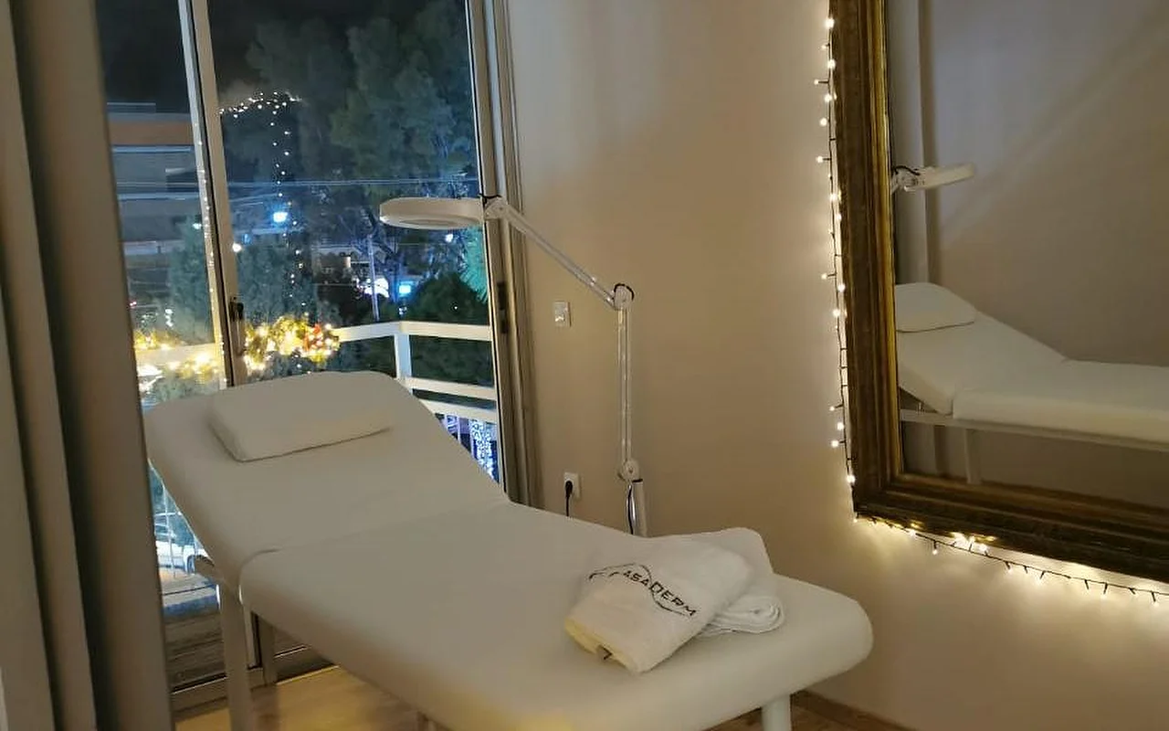 Una sala de tratamiento con iluminación suave, una cama blanca con una toalla con el logotipo de CasaDerm, una lupa profesional, luces de hilo enmarcando un espejo dorado y un ventanal que da al paisaje nocturno de las calles de Glyfada.