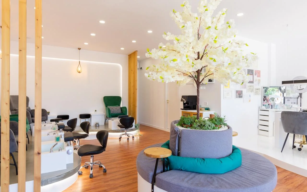 Un salón de uñas abierto con inspiración natural, con un gran árbol decorativo de flores blancas como pieza central, butacas de terciopelo verde esmeralda en las estaciones de pedicura, divisores de listones de madera y suelo de madera cálida.