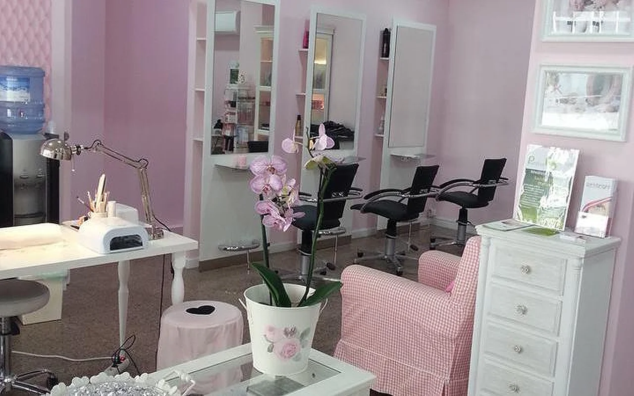 El interior de un luminoso salón de peluquería y uñas con temática en rosa. A la izquierda, una estación de manicura blanca con flexo y lámpara de curado de uñas. En el centro, una orquídea en una maceta blanca sobre una mesa de centro junto a un sillón de cuadros vichy en rosa y blanco. Al fondo, varias estaciones de peluquería con espejos altos y sillones de salón negros contra paredes rosa claro.