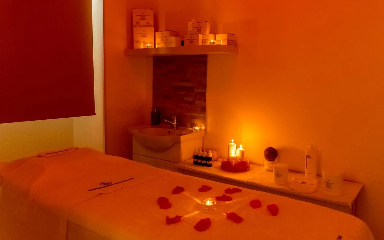 Una sala de masaje con una iluminación cálida y envolvente en tonos anaranjados y velas encendidas. Pétalos de rosa rojos dispuestos en círculo alrededor de una vela central sobre la camilla de tratamiento, con productos de belleza ordenados en una estantería detrás.