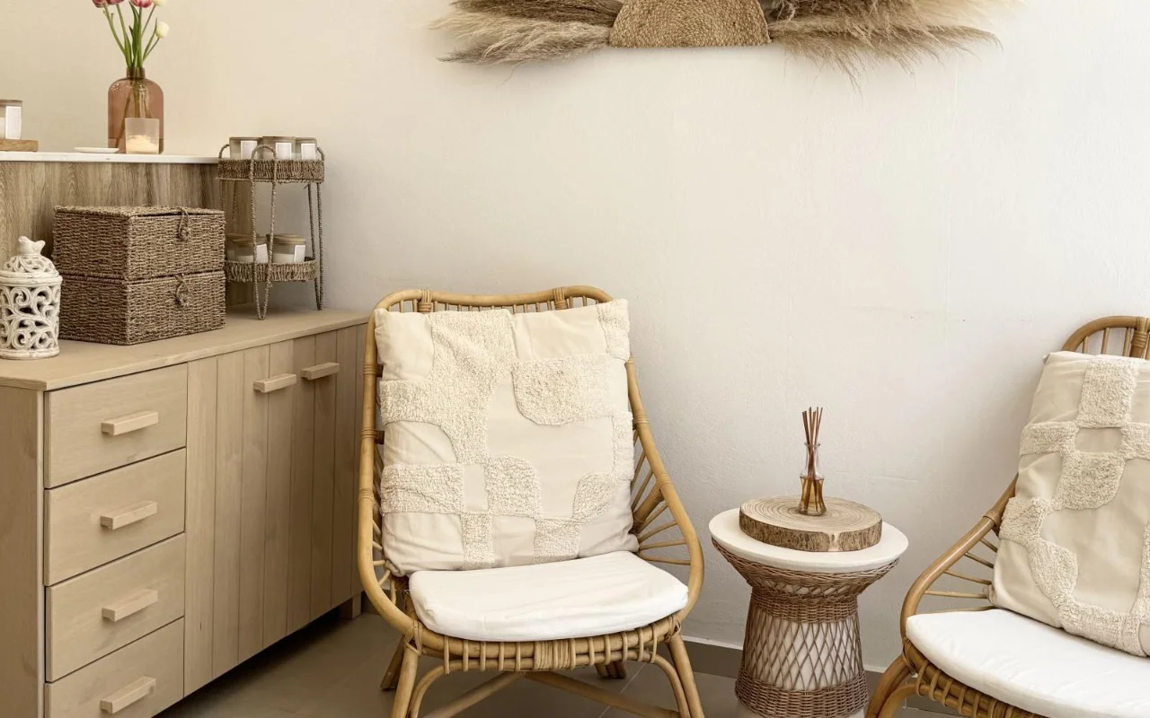 Una zona de espera relajante con butacas de ratán, cojines de textura crema, una mesa auxiliar de mimbre con un difusor de ambientador, cestas de almacenaje tejidas y hierba de la pampa seca en la pared sobre armarios de madera clara.