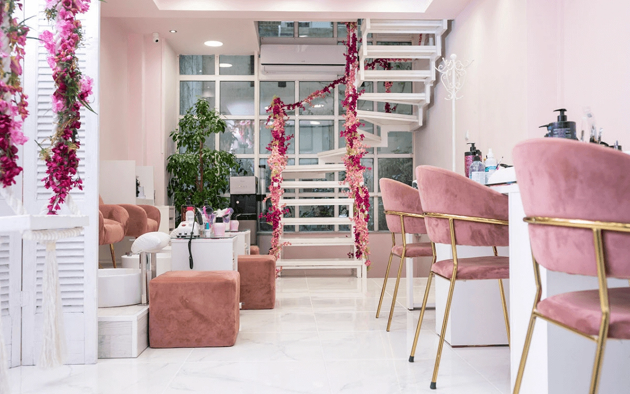 Un salón de uñas femenino con sillones de terciopelo rosa empolvado, una escalera de caracol blanca decorada con flores artificiales rosas, biombos con contraventanas, y estaciones de manicura con patas doradas sobre suelo efecto mármol.