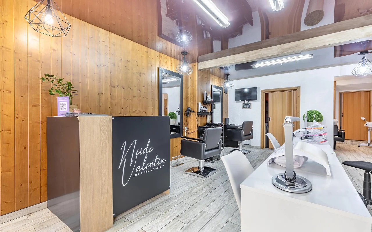 Un salón de belleza contemporáneo con mostrador de recepción a la izquierda rotulado con 'Neide Valentim Instituto de Beleza'. Las paredes están revestidas con paneles verticales de madera cálida y grandes espejos. Sillones de peluquería negros frente a los espejos, y una estación de manicura blanca con sillas blancas a lo largo del lado derecho del suelo de baldosas grises.
