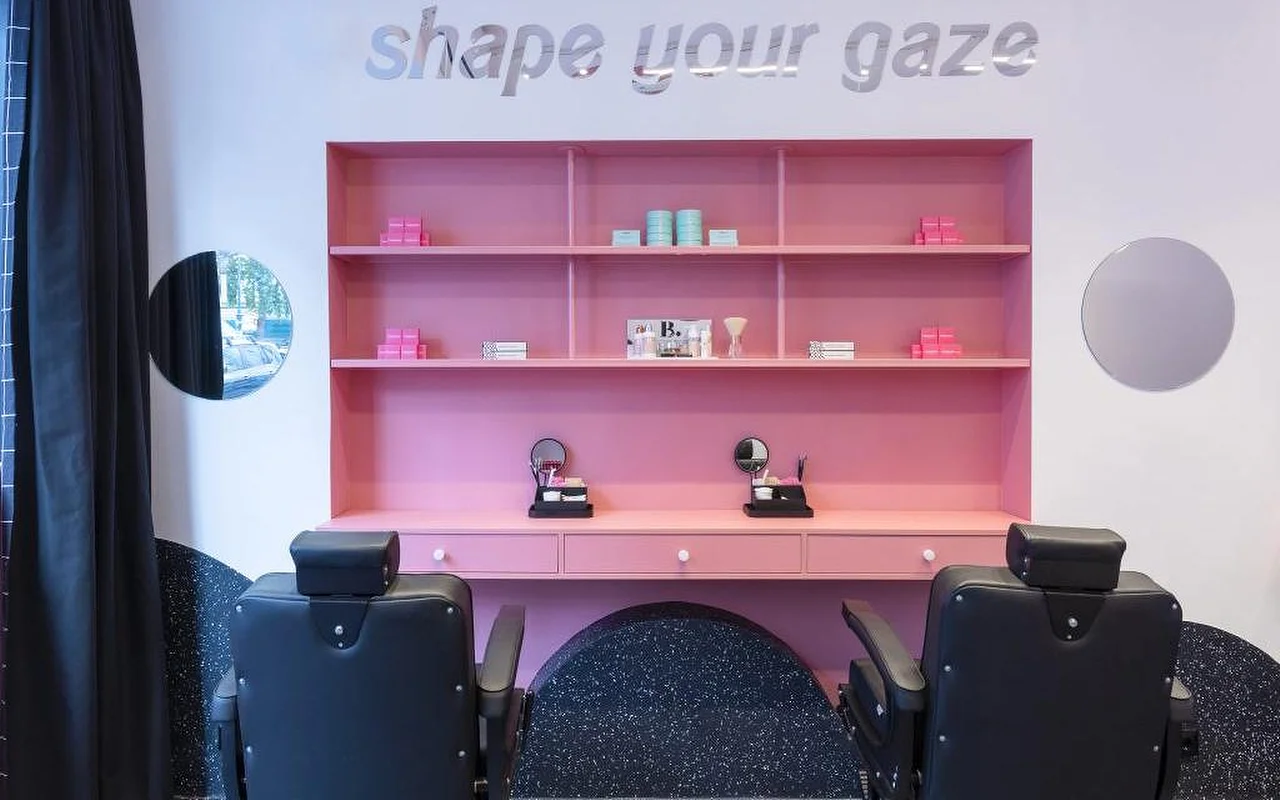Un estudio especializado en cejas y pestañas con una estantería rosa llamativa con productos de belleza, letras plateadas que dicen 'shape your gaze' en la pared, y sillones de tratamiento en cuero negro sobre un suelo oscuro moteado.