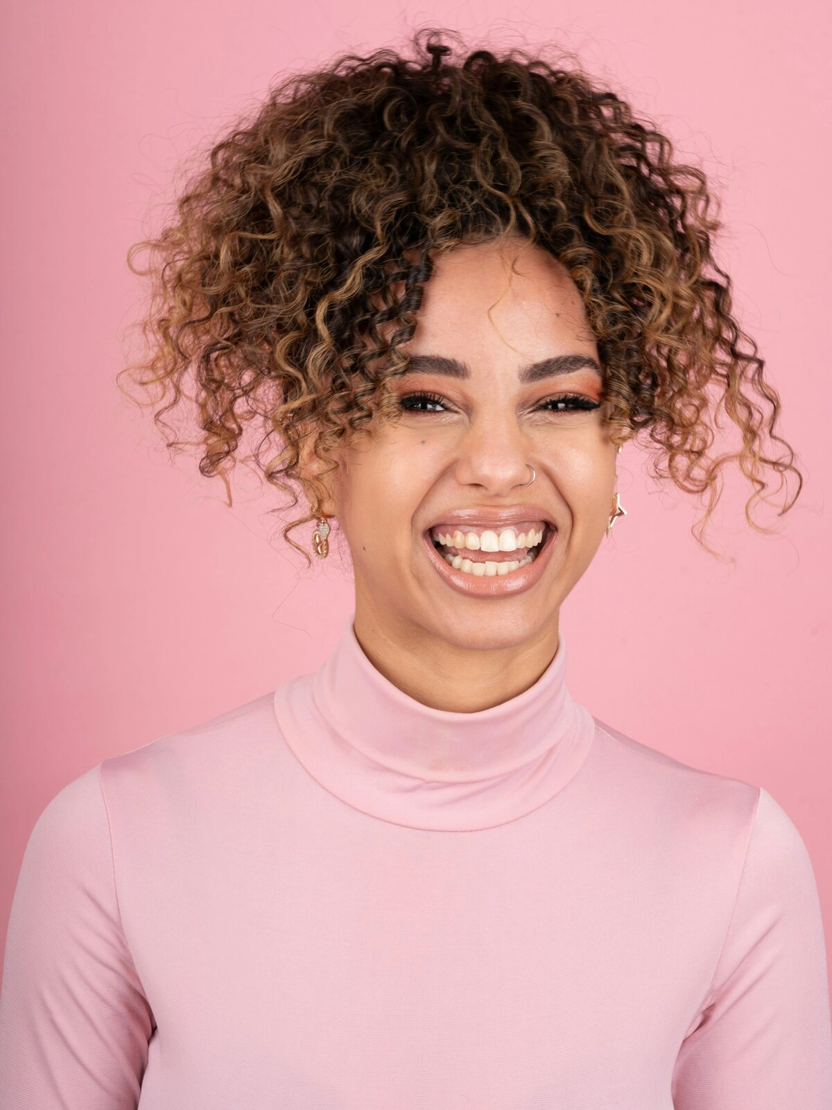 Foto de estudio de una mujer sonriente con balayage sobre pelo afro, vestida con un jersey de cuello alto rosa suave