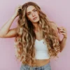 Foto de estudio de una mujer con el pelo larguísimo, ondulado y besado por el sol con balayage, vestida con vaqueros azul claro y un top corto blanco de punto