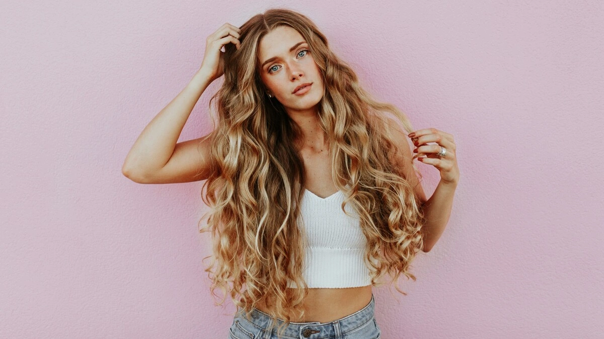 Foto de estudio de una mujer con el pelo larguísimo, ondulado y besado por el sol con balayage, vestida con vaqueros azul claro y un top corto blanco de punto