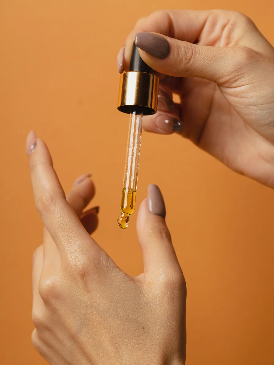 Primer plano de dos manos con manicura; una de ellas sostiene un cuentagotas de cristal del que cae aceite de cutícula