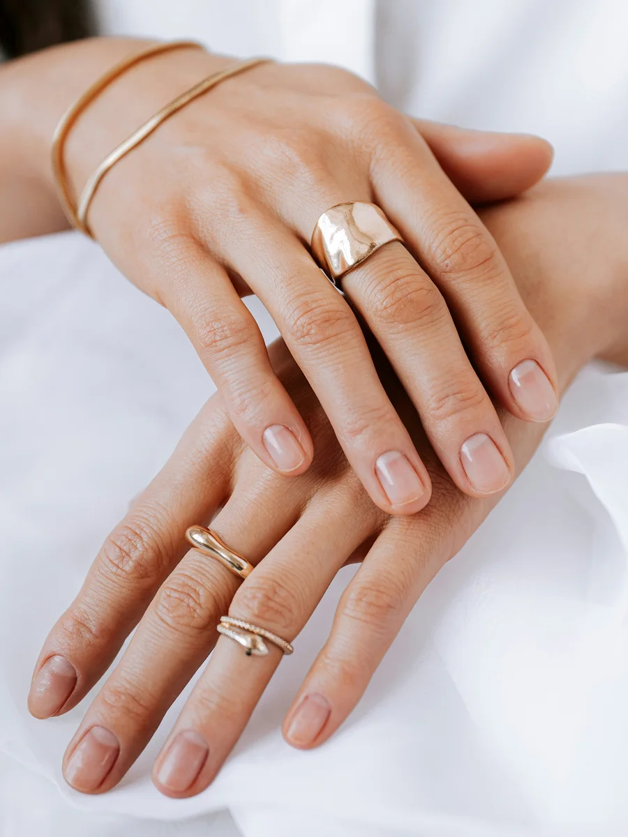 Primer plano de dos manos apoyadas sobre una sábana blanca que muestran unas uñas naturales y sanas, adornadas con anillos y pulseras de oro rosa