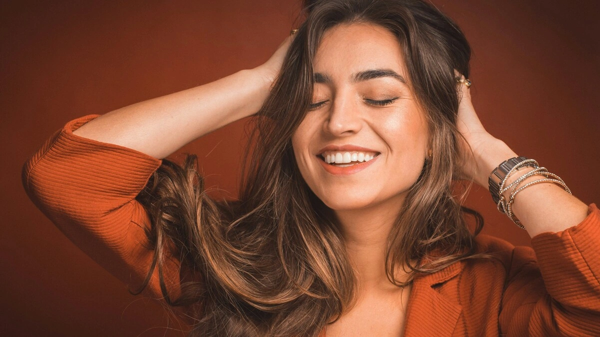 Primer plano de una mujer sonriendo con los ojos cerrados mientras pasa las manos por su pelo castaño, largo y suave
