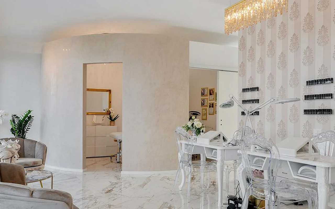 El elegante interior de MC Beauty Center con suelo de efecto mármol, paredes curvas blancas y una lámpara de araña de cristal dorada. A la derecha hay una estación de manicura con sillas transparentes, mientras que unos sillones de terciopelo y una puerta con marco dorado dan paso a una cabina de tratamiento al fondo.