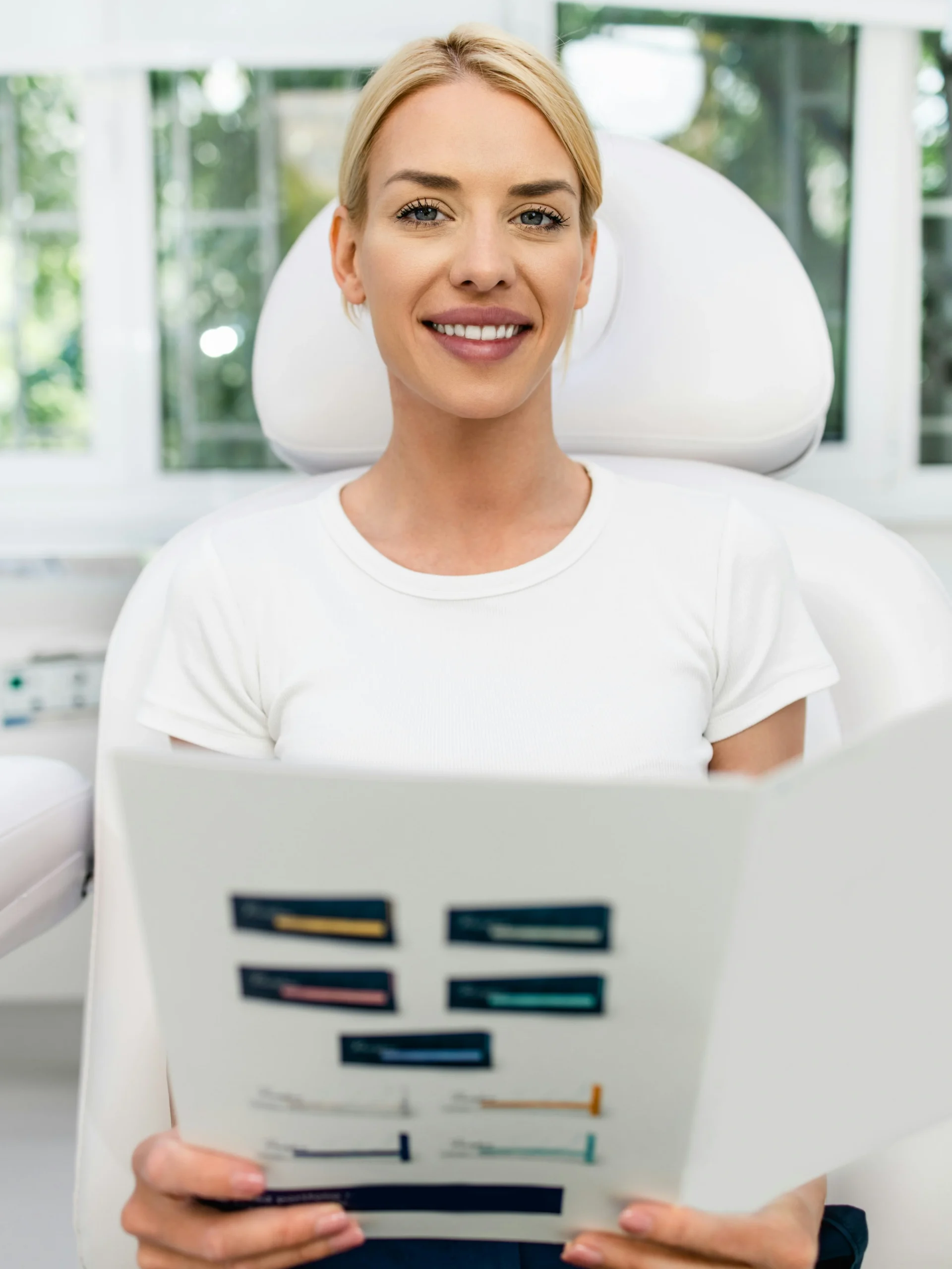Una clienta sentada en una clínica luminosa y limpia sonríe mientras sostiene un folleto informativo sobre tratamientos faciales