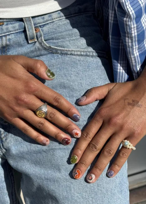Manicura todo vale en uñas cortas, cada uña de un color y un diseño distintos, con una estrella roja y una voluta naranja, apoyada sobre unos vaqueros.