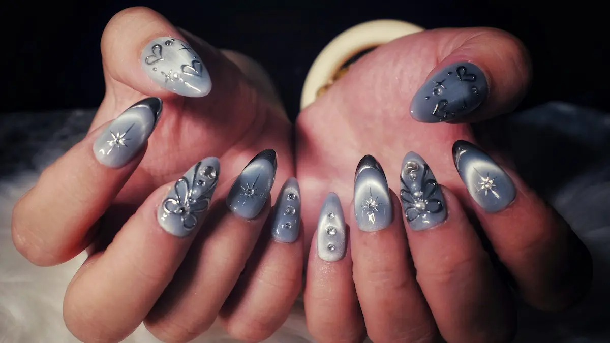 Primer plano de las manos de una mujer con uñas acrílicas elaboradas: diseño cat-eye gris, corazones y estrellas plateados y piedras brillantes