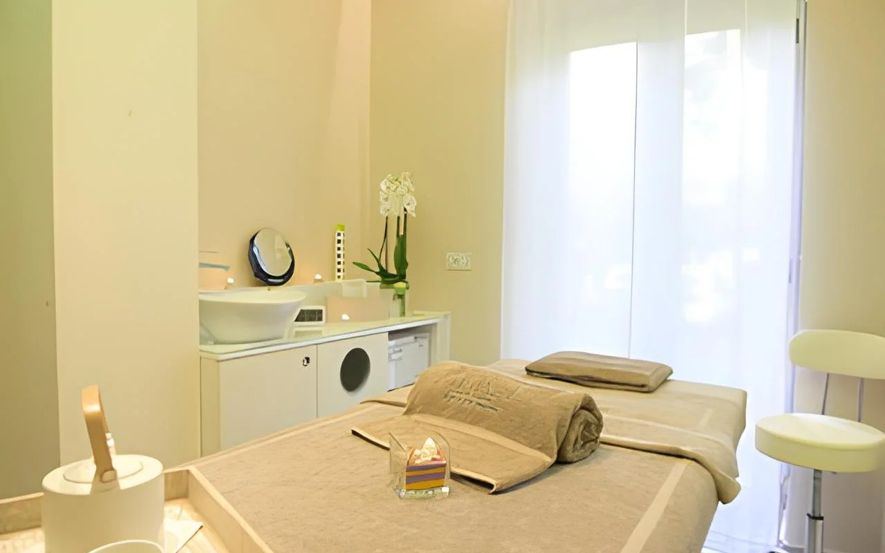 Una cabina de tratamiento serena en Alice Beauty Clinique, con una camilla vestida con ropa de cama en tonos topo suaves, orquídeas blancas sobre la encimera, velas junto al lavabo y cortinas vaporosas que filtran la luz natural a través de un gran ventanal.