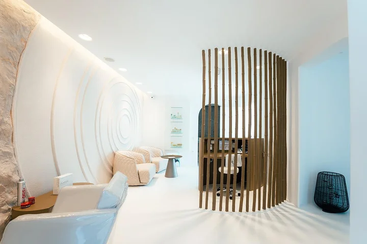 Un interior de spa tranquilo con aire de gruta, de paredes blancas curvas, una de ellas tallada en ondas concéntricas, con iluminación indirecta suave, sillones acolchados color crema y un biombo curvo de listones de madera que delimita un rincón de tratamiento tranquilo.