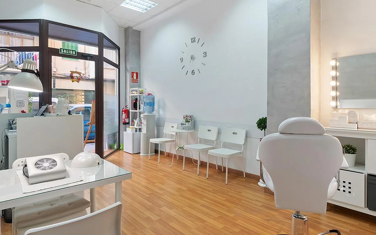 Un salón luminoso y blanco con suelo de madera clara, una fila de sillas de espera blancas, un reloj de pared minimalista, un espejo de tocador iluminado, una camilla reclinable blanca y una estación de manicura junto a la ventana.