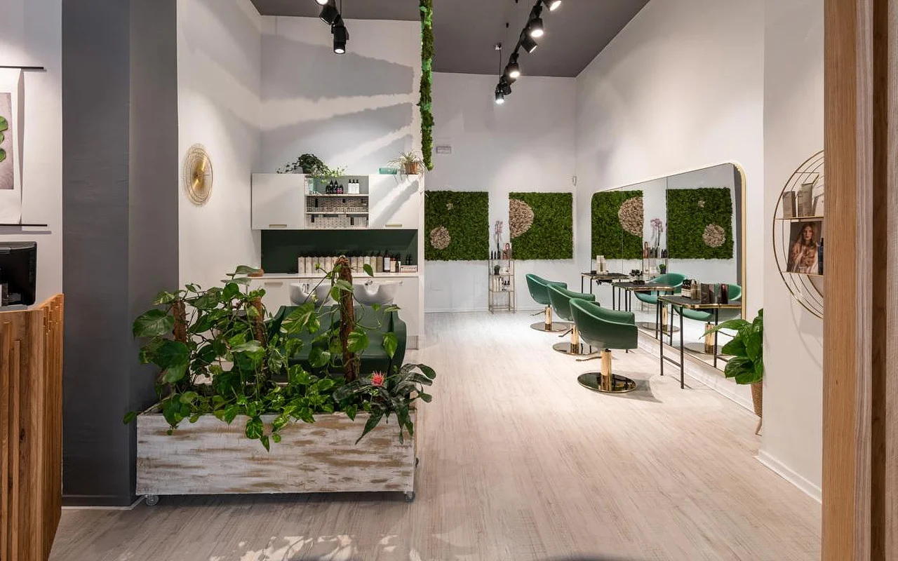 Una peluquería llena de plantas con suelos de madera clara, una vieja jardinera blanca rebosante de vegetación, arte mural de musgo verde, sillones de peinado de terciopelo verde esmeralda con detalles dorados y un lavacabezas al fondo.