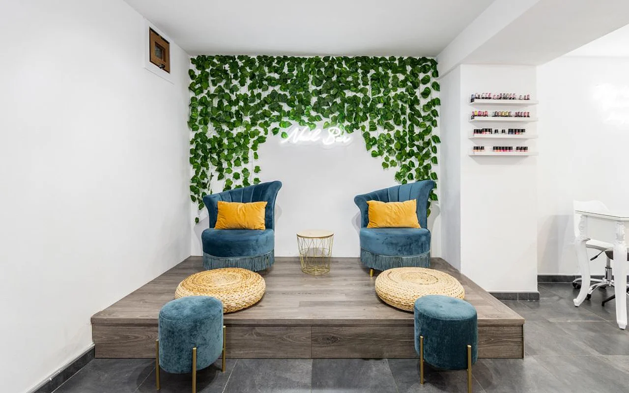 Un rincón de centro de uñas con una pared de follaje verde en cascada y un pequeño cartel de neón, dos butacas de terciopelo turquesa con cojines mostaza sobre una tarima de madera, pufs de mimbre a modo de reposapiés y estanterías con esmaltes de colores a la derecha.