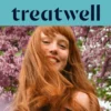 Primer plano de una mujer joven pelirroja y sonriente con flequillo, con el cabello moviéndose al viento frente a un fondo de flores de lila rosas. En la parte superior, una franja azul claro muestra el logotipo de "Treatwell" en azul oscuro.