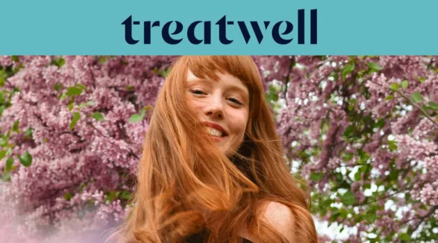 Primer plano de una mujer joven pelirroja y sonriente con flequillo, con el cabello moviéndose al viento frente a un fondo de flores de lila rosas. En la parte superior, una franja azul claro muestra el logotipo de "Treatwell" en azul oscuro.