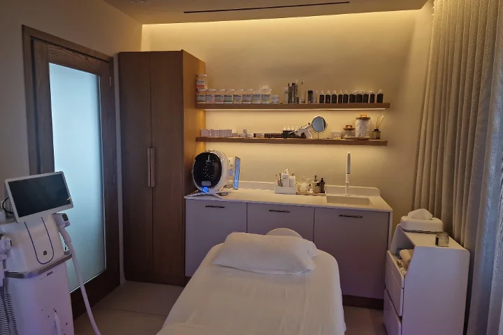 Una cabina de tratamiento de luz cálida, con una camilla facial blanca y toallas dobladas con esmero, estanterías de madera oscura llenas de productos para la piel, una pantalla de análisis de la piel y aparatos de tratamiento al lado, clínica pero acogedora.