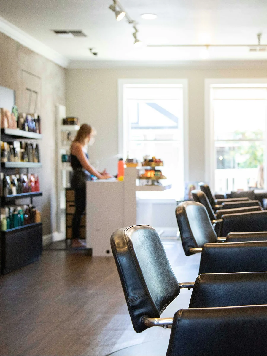 Imagen del interior de una peluquería limpia, luminosa y bien ventilada, con grandes ventanales, sillones negros y estanterías con productos