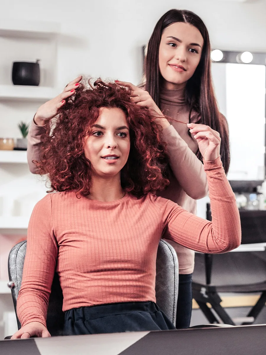 Una clienta con el pelo rizado teñido de rojo, sentada en la peluquería mientras su estilista le explica el tratamiento