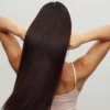 Vista de espaldas de una mujer con un top blanco de tirantes y los brazos en alto, pasando los dedos por su larga y brillante melena castaña