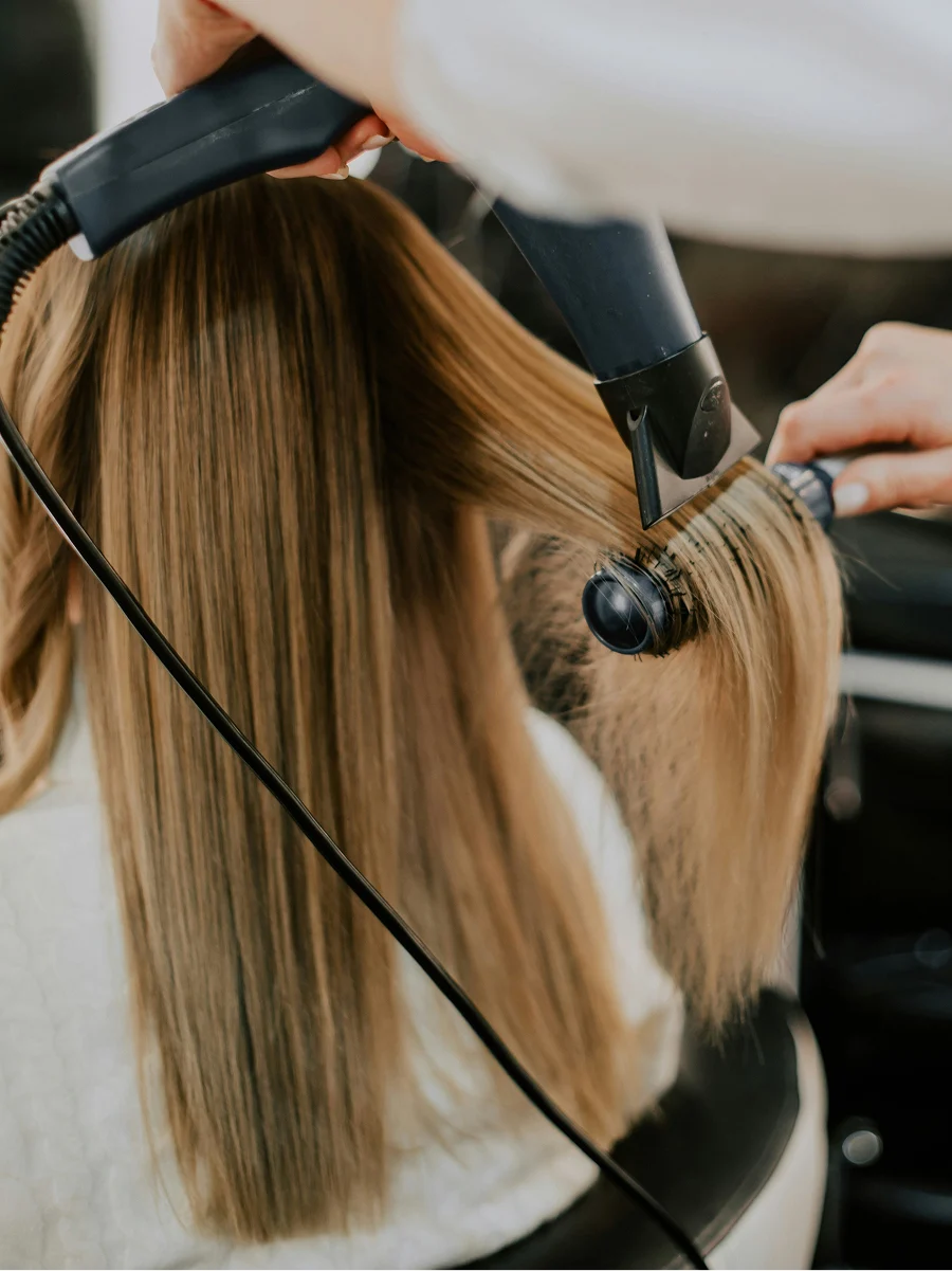 Vista de espaldas de una mujer en la peluquería mientras una estilista le seca el pelo liso durante una sesión de tratamiento de queratina