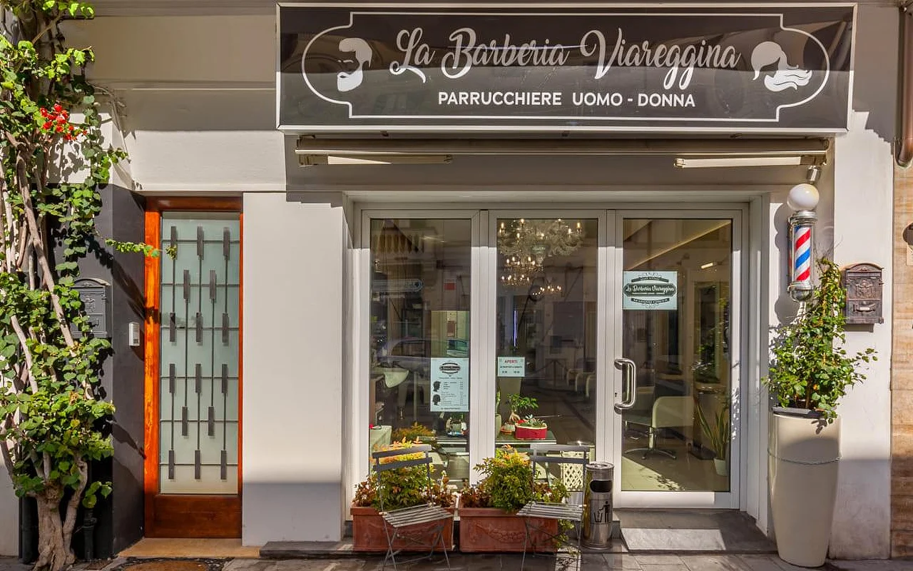 El escaparate de La Barberia Viareggina en un día soleado, con un clásico poste de barbero rojo, blanco y azul a la derecha de las puertas de cristal. Plantas en macetas y enredaderas enmarcan la entrada, y por las ventanas se ve una lámpara de araña en el interior.