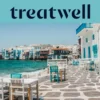 El paseo marítimo de la Pequeña Venecia en Mykonos: casas encaladas con balcones de madera de colores al borde del agua, el mar Egeo turquesa al fondo y, en primer plano, mesas de café blancas y verde azulado dispuestas sobre una terraza adoquinada.