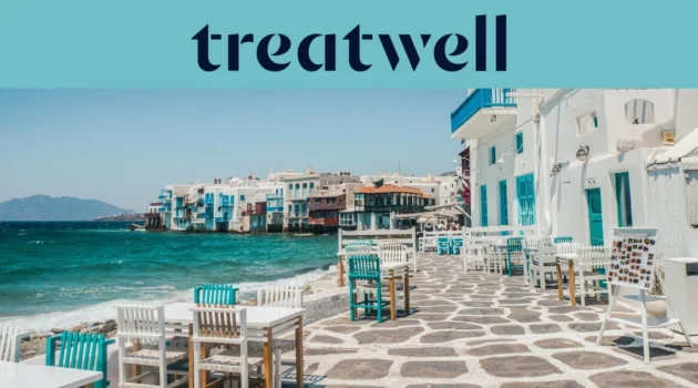 El paseo marítimo de la Pequeña Venecia en Mykonos: casas encaladas con balcones de madera de colores al borde del agua, el mar Egeo turquesa al fondo y, en primer plano, mesas de café blancas y verde azulado dispuestas sobre una terraza adoquinada.