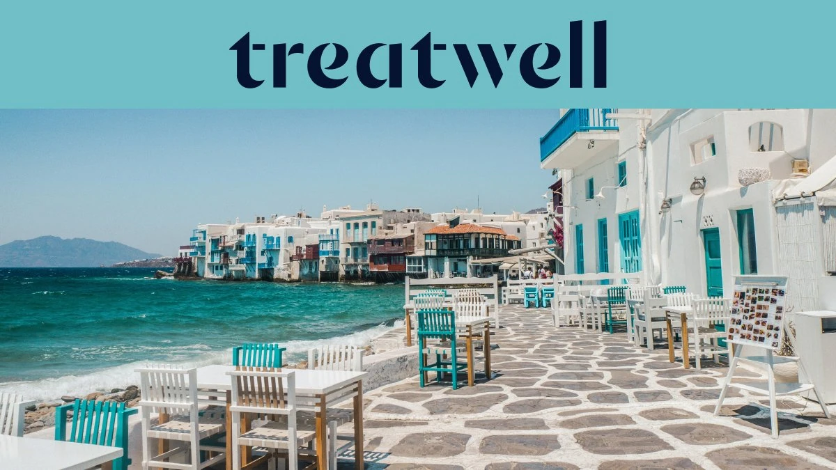 El paseo marítimo de la Pequeña Venecia en Mykonos: casas encaladas con balcones de madera de colores al borde del agua, el mar Egeo turquesa al fondo y, en primer plano, mesas de café blancas y verde azulado dispuestas sobre una terraza adoquinada.
