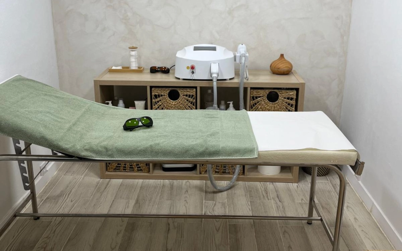 Una sala de tratamientos en tonos neutros con una camilla de estructura metálica cubierta con una toalla verde salvia, un par de gafas protectoras oscuras encima, un aparato de láser sobre una consola de madera y cestas de mimbre debajo.