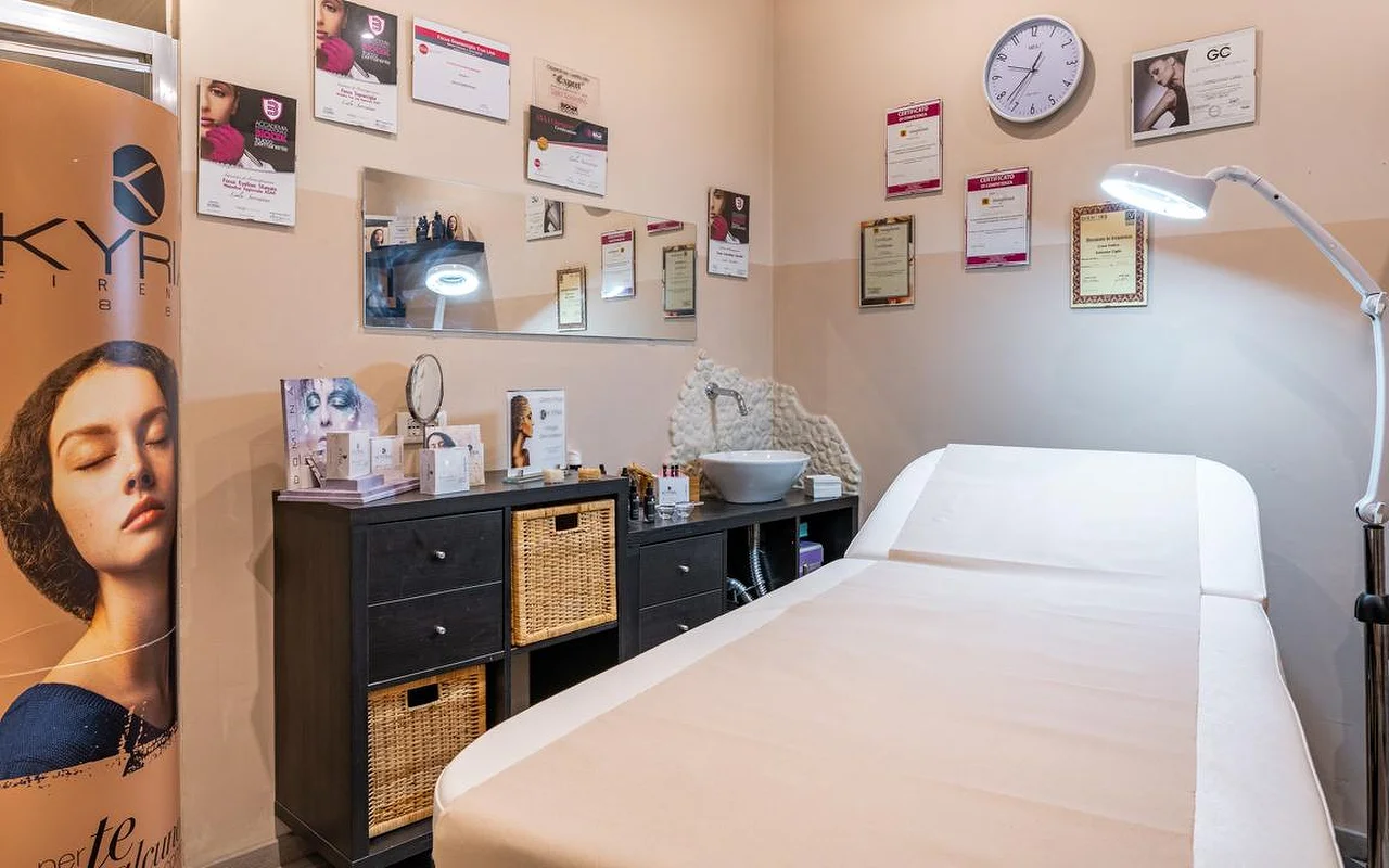 Una cabina de tratamiento en Oltre la bellezza con una camilla blanca, una lámpara lupa regulable y paredes cubiertas de certificados y diplomas profesionales. Bajo un espejo hay muebles de almacenaje de mimbre oscuro, con productos de cuidado de la piel colocados sobre la encimera.