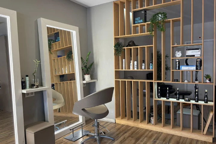 Un puesto de peluquería con un sillón gris frente a un espejo enmarcado e iluminado con LED, un panel-estantería de listones de madera con productos de peinado, un reloj y plantas, sobre un suelo de efecto madera.