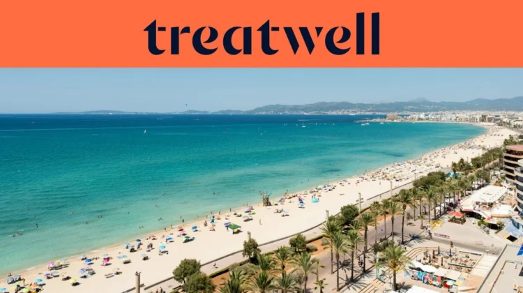 Vista aérea de la Playa de Palma en Mallorca, con una larga curva de arena dorada, hileras de sombrillas, agua turquesa en calma y un paseo bordeado de palmeras junto a la ciudad, con colinas a lo lejos bajo un cielo azul despejado.
